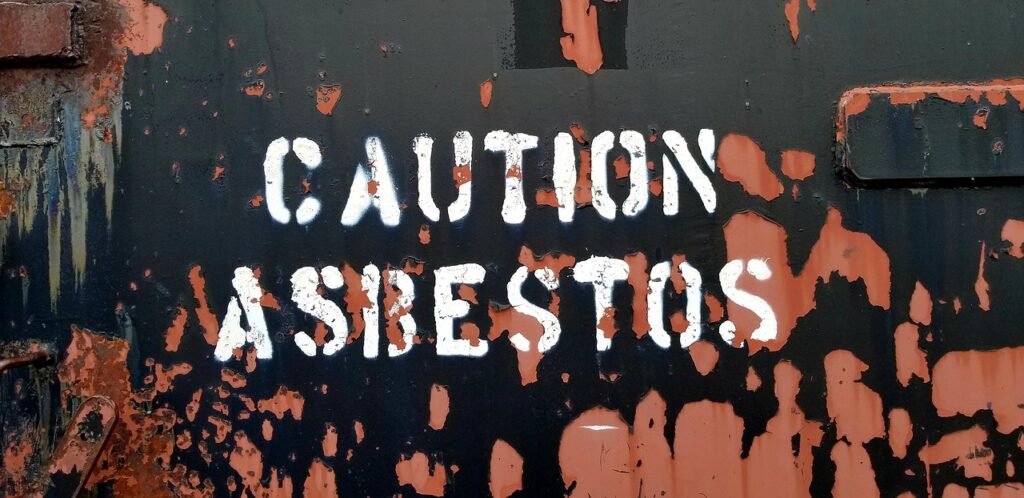 Close up of a weathered metal surface with peeling orange paint and white stencil text reading Caution Asbestos