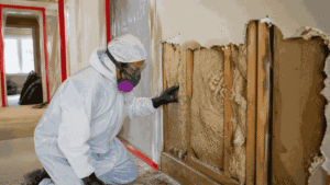 Worker in hazmat suit and respirator checking exposed insulation and wall studs in a contained indoor remediation site.